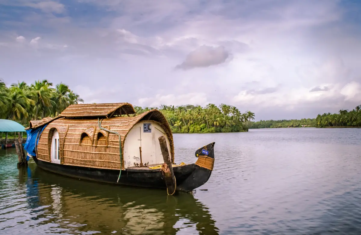 Wooden kettuvallam houseboat moored at a palm-covered bank under a cloudy evening sky